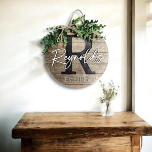 May include: A round wooden sign with a black letter "R" and the name "Reynolds" written in white script. The sign also includes the text "Est. 2019" and is decorated with a green leafy garland.