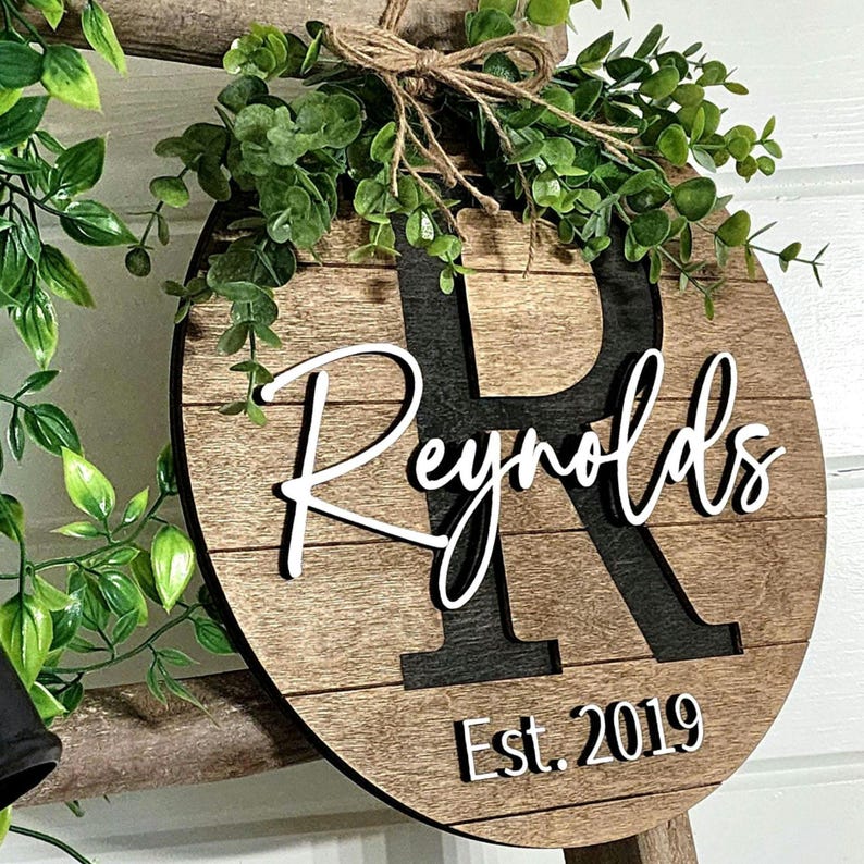 May include: A round wooden sign with a faux wood grain finish. The sign features a large black letter "R" with a white outline. The name "Reynolds" is written in white cursive script above the letter "R". The text "Est. 2019" is written in white below the letter "R". The sign is decorated with a green leafy garland.