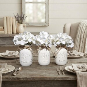 May include: Three white painted glass Ball mason jars, each filled with white hydrangea flowers. Each jar is wrapped with brown twine around the neck. The jars are arranged on a wooden table with place settings including plates and silverware.