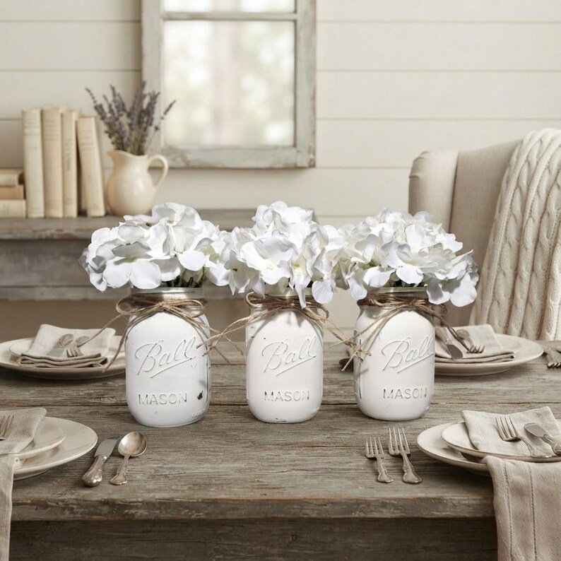 May include: Three white painted glass Ball mason jars, each filled with white hydrangea flowers. Each jar is wrapped with brown twine around the neck. The jars are arranged on a wooden table with place settings including plates and silverware.