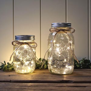 May include: Two clear glass Mason jars with silver lids, filled with fairy lights. Each jar is adorned with a brown twine bow. The jars are on a wooden surface, with greenery in the background, creating a warm, inviting ambiance.