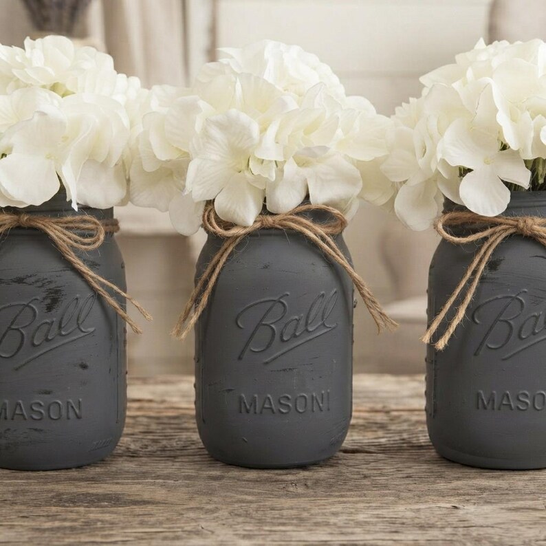 May include: Three matte charcoal gray glass mason jars, each embossed with the word Ball and Mason, are filled with white hydrangea flowers. Each jar is tied with a natural brown jute twine bow around the neck, featuring a distressed finish.