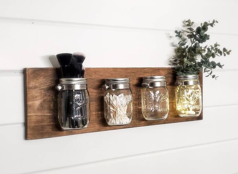 May include: Four glass jars with metal lids are mounted on a wooden shelf. The jars are filled with various items, including makeup brushes, cotton swabs, and string lights. The shelf is mounted on a white wall.