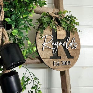May include: A round wooden sign with the last name "Reynolds" in white script lettering and a large black "R" in the center. The sign also includes the text "Est. 2019" in white lettering. The sign is hanging on a wooden ladder with greenery around it.