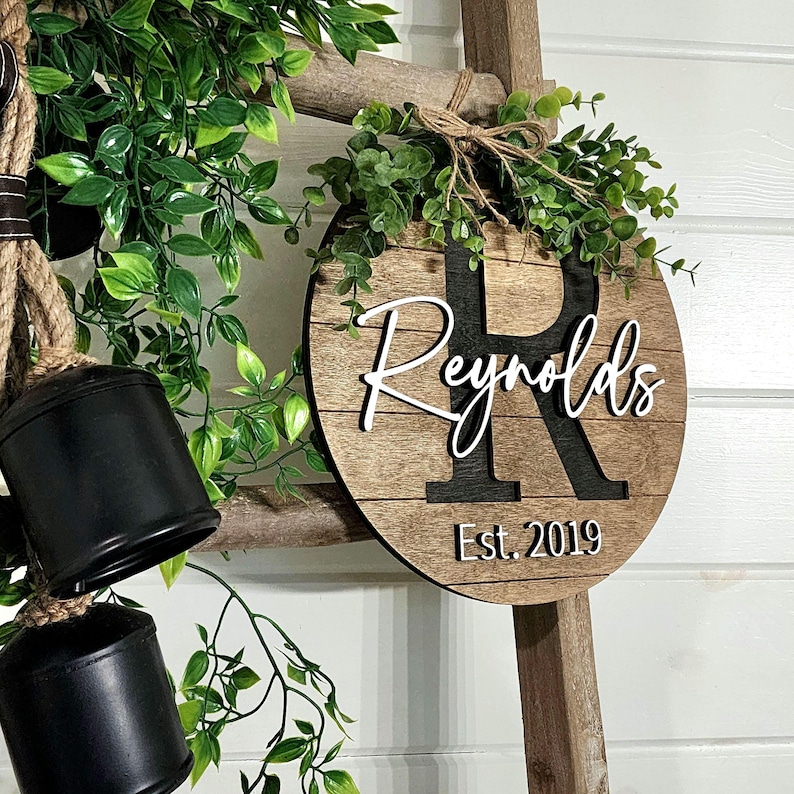 May include: A round wooden sign with the last name "Reynolds" in white script lettering and a large black "R" in the center. The sign also includes the text "Est. 2019" in white lettering. The sign is hanging on a wooden ladder with greenery around it.