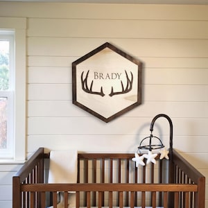 May include: A wooden sign with the name "BRADY" and deer antlers is mounted on a wall above a crib. The sign is a light wood color with a dark brown border. A baby mobile with white star-shaped ornaments hangs above the crib.