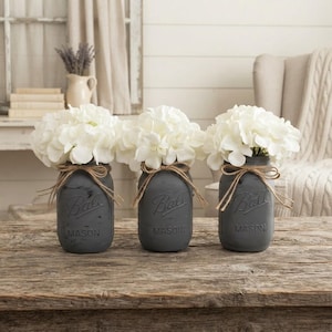 May include: Three matte charcoal gray glass Ball Mason jars, each filled with a bouquet of white hydrangea flowers. Each jar features a natural jute twine bow tied around the neck, with the embossed text Ball and Mason visible on the front.