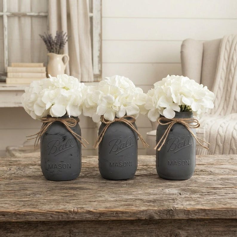 May include: Three matte charcoal gray glass Ball Mason jars, each filled with a bouquet of white hydrangea flowers. Each jar features a natural jute twine bow tied around the neck, with the embossed text Ball and Mason visible on the front.