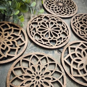 May include: Six wooden coasters with intricate laser-cut mandala designs. The coasters are circular and feature a variety of patterns, including floral and geometric motifs. The coasters are made of a dark brown wood and have a natural finish.
