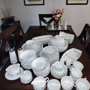 May include: A large collection of white ceramic dinnerware with a blue and gray geometric pattern. The set includes plates, bowls, cups, serving dishes, and a covered casserole dish. A salt and pepper shaker set is also included.