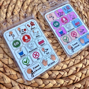 May include: Two communication boards with clear plastic sleeves, featuring colorful icons and text. The boards include symbols for "Go," "Stop," "Drink," "Restroom," and "Eat." The text includes "Want," "More," "Help," "Yes," "No," "I need a break," and "All done."