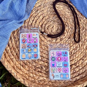 May include: Two communication boards with colorful icons and text, designed for nonverbal communication. The boards are in clear plastic holders with black lanyards. The boards are on a woven wicker surface with a blue fabric in the background.