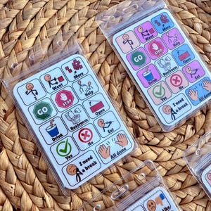 May include: Several communication boards with colorful icons and text, including "Want," "More," "Help," "Go," "Drink," and "Restroom." Each board is encased in a clear plastic sleeve, designed for visual communication.