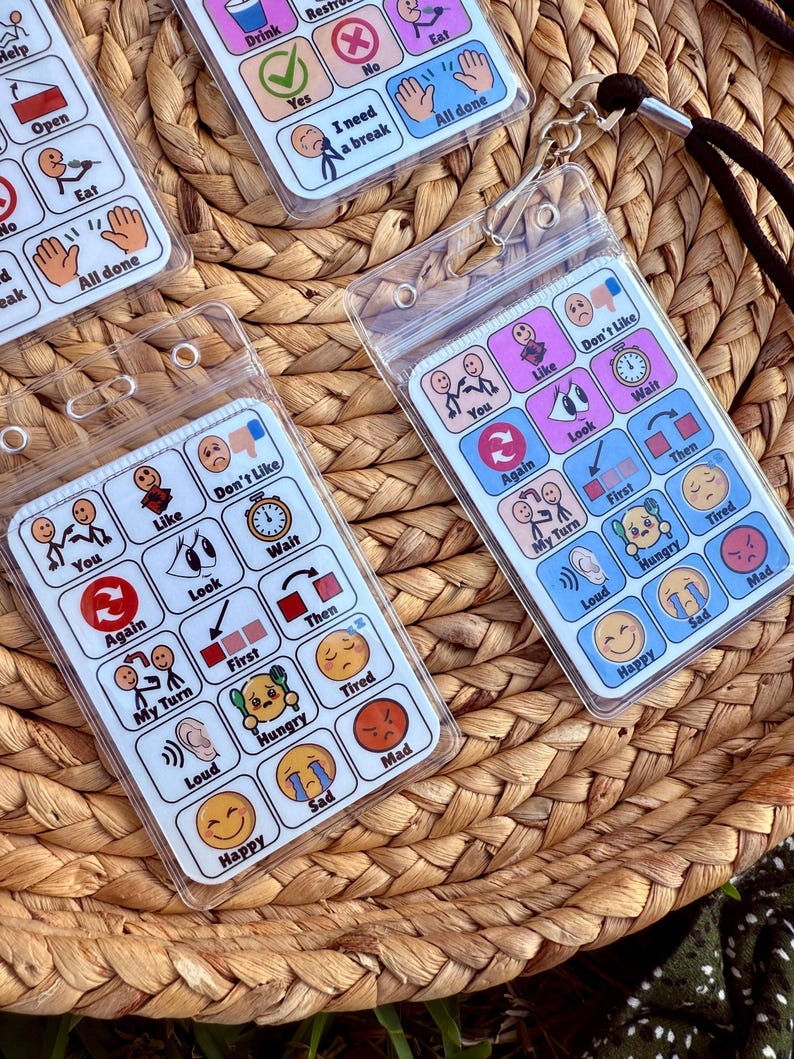 May include: Clear plastic communication boards with various icons and phrases, including "Eat," "Drink," and emotional expressions. Each board is designed to be worn around the neck with a black lanyard. The boards are displayed on a woven basket.