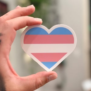May include: A heart-shaped sticker featuring the transgender pride flag design. The sticker displays horizontal stripes in light blue, pink, and white, arranged in a symmetrical pattern with a white stripe in the center, flanked by pink and blue.