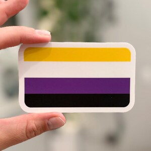 May include: A rectangular sticker featuring the non-binary pride flag, which consists of four horizontal stripes in yellow, white, purple, and black. The sticker has rounded corners and a white border surrounding the colorful flag design.