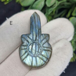 May include: A carved labradorite guitar pendant. The pendant is a mix of blue, green, and grey hues. The guitar design features a sunburst pattern on the body and vertical lines representing the strings. The top of the pendant has a rectangular shape.