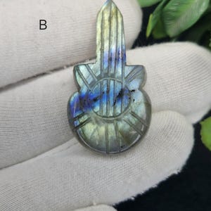 May include: A carved labradorite gemstone pendant in the shape of a guitar. The pendant features a mix of blue, green, and grey hues with carved details. The gemstone is held in a gloved hand against a white background.