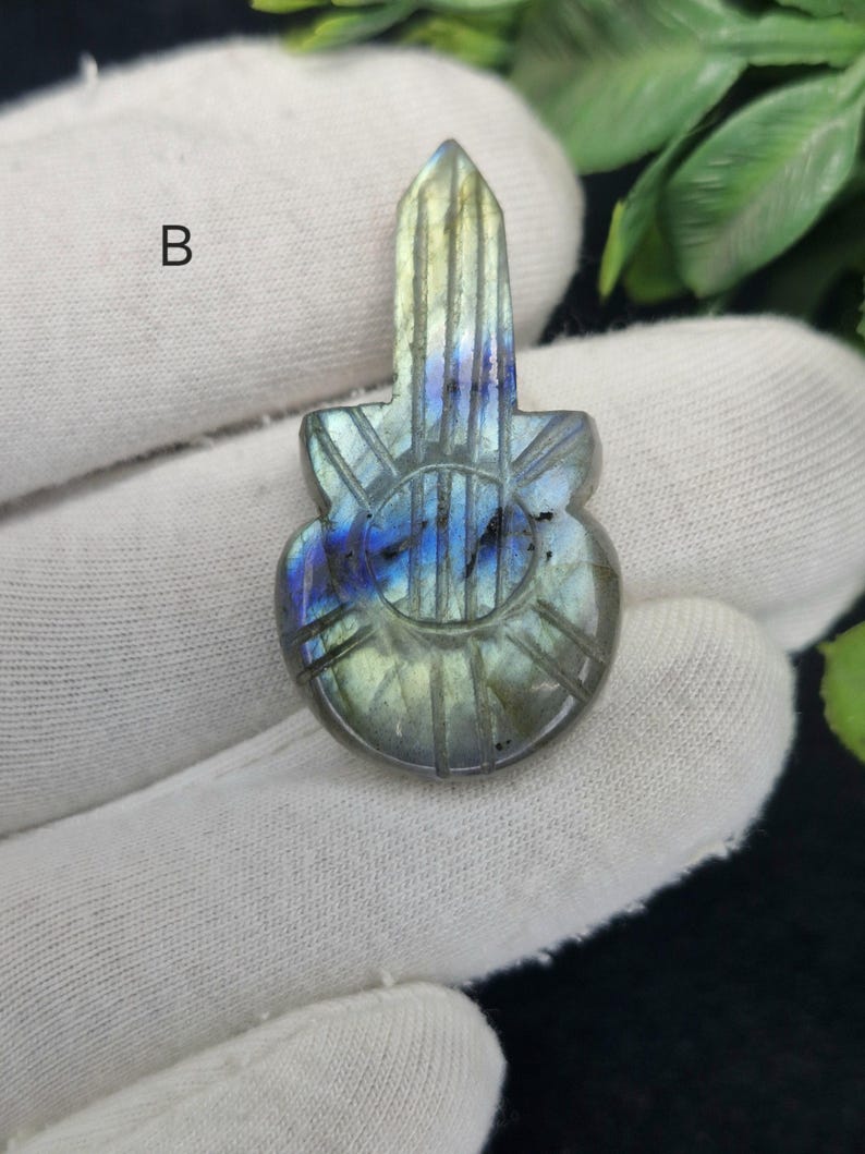 May include: A carved labradorite gemstone pendant in the shape of a guitar. The pendant features a mix of blue, green, and grey hues with carved details. The gemstone is held in a gloved hand against a white background.