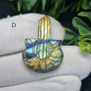May include: A labradorite pendant carved in a guitar shape. The gemstone shows blue, green, and gold hues. The letter "D" is visible in the upper left corner. The pendant is displayed against a white fabric.