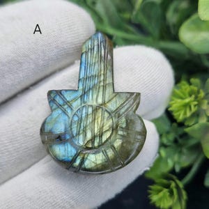 May include: A carved labradorite gemstone pendant in a guitar shape. The stone has iridescent blue, green, and gold colours. The pendant features etched lines and a circular design in the centre.