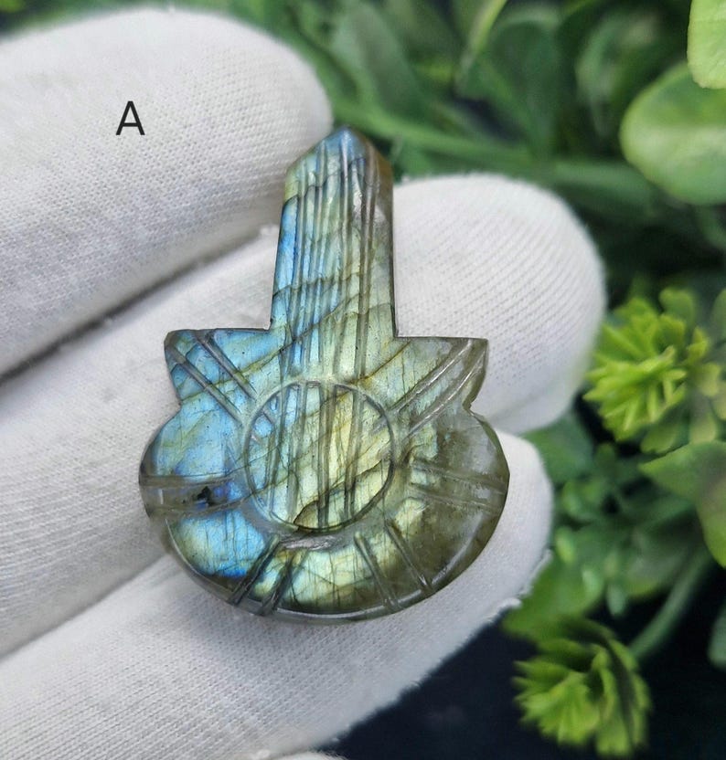 May include: A carved labradorite gemstone pendant in a guitar shape. The stone has iridescent blue, green, and gold colours. The pendant features etched lines and a circular design in the centre.