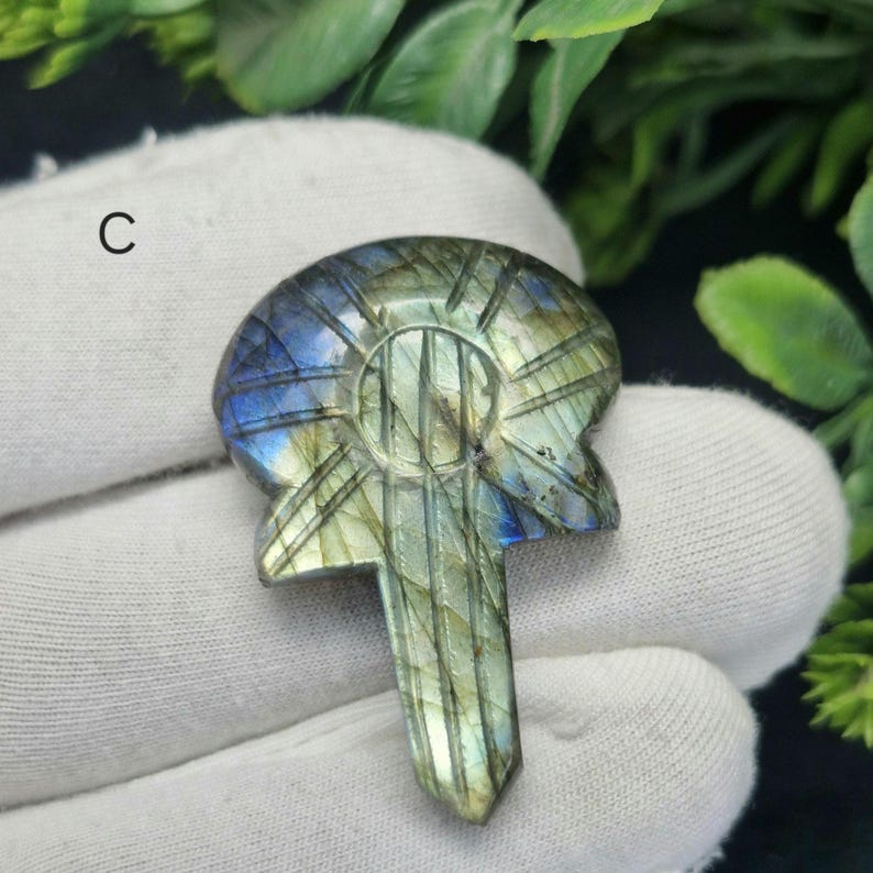 May include: A carved labradorite gemstone key pendant. The pendant has a key design with a rounded top and a pointed bottom. The stone has blue, green, and grey hues with etched lines.