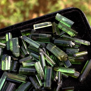 Op de afbeelding: Een close-up van een verzameling ruwe, groene toermalijnkristallen. De kristallen zijn van verschillende groottes en vormen, met een doorschijnend, glasachtig uiterlijk. Ze worden weergegeven op een zwarte ondergrond, met een vage groene achtergrond.