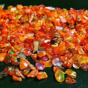 May include: A large pile of small, orange, red, and iridescent opal chips. The chips are scattered on a black surface.