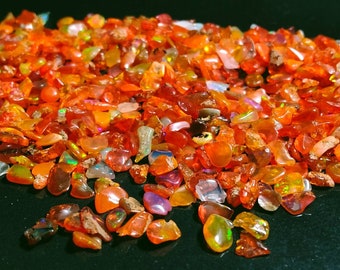 May include: A large pile of small, orange, red, and iridescent opal chips. The chips are scattered on a black surface.