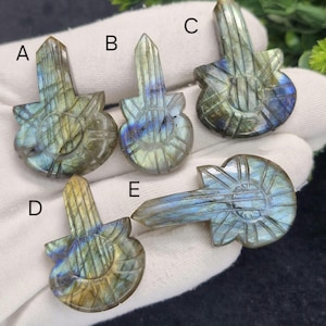 May include: Five carved labradorite guitar pendants. Each pendant has a detailed guitar shape with a rectangular top and a rounded body. The stones display iridescent blue, green, and gold hues. The pendants are displayed on a white surface.