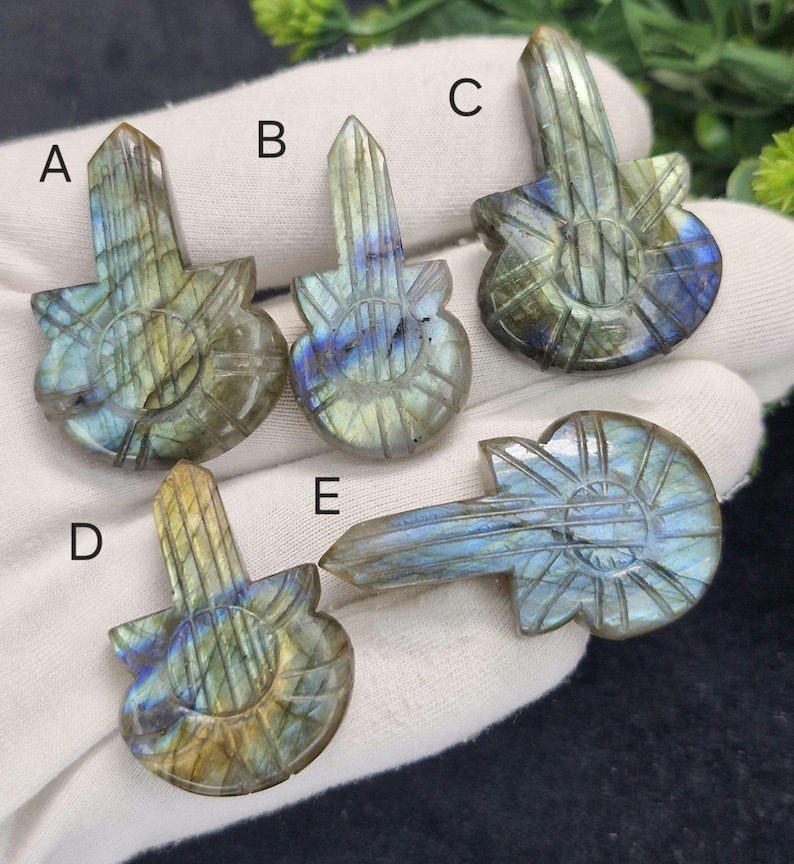 May include: Five carved labradorite guitar pendants. Each pendant has a detailed guitar shape with a rectangular top and a rounded body. The stones display iridescent blue, green, and gold hues. The pendants are displayed on a white surface.