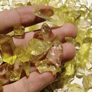 May include: A handful of translucent, yellow-green crystals. The crystals vary in size and shape, with some appearing more rectangular and others irregular. The light shines through the crystals, highlighting their clarity and color. The crystals are held in a hand.