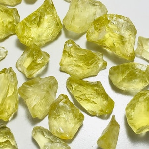 May include: A collection of rough-cut, translucent yellow gemstones. The stones vary in size and shape, with a crystalline appearance. The color ranges from pale to vibrant yellow, with visible internal structures. The gemstones are scattered across a white surface.