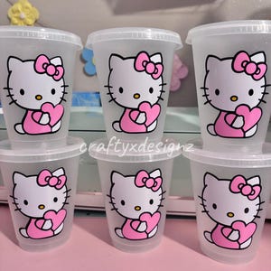 May include: Six translucent plastic cups with clear lids, each featuring a graphic of a white cat character with a pink bow holding a pink heart. The cups are arranged in two rows of three, and the text craftyxdesignz is printed in white.