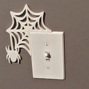 Könnte beinhalten: Ein weißer Lichtschalter mit einem ausgeschnittenen Spinnennetz- und Spinnenmotiv. Der Schalter ist an einer Wand montiert.