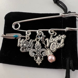 May include: A silver-toned safety pin with dangling charms. The charms include the words "Wolf Girl" and "Vampire Girl", a fleur-de-lis, a feather with a turquoise accent, and a pink pearl. The pin is displayed on a black velvet pouch.