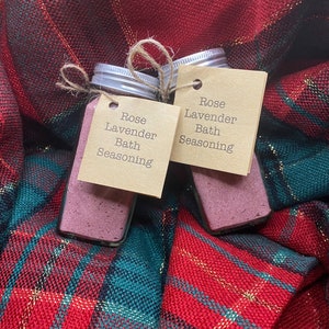 May include: Two small glass jars filled with pink powder, labeled "Rose Lavender Bath Seasoning," are tied with twine. The jars sit on a red, green, and gold plaid fabric. The jars have silver lids and small brown tags.