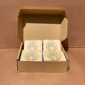 May include: A cardboard box with four bars of goat's milk soap. The soap bars are labeled "Copper Rabbit Crafts" and have different scents: Honey Lavender Oat, Lemon Verbena Basil, Fresh Pine, and Lemon Poppy Seed.