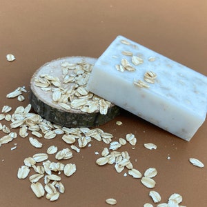 May include: Two bars of white soap with oatmeal embedded in the soap. The soap is on a wooden slice with oatmeal scattered around it on a brown surface.