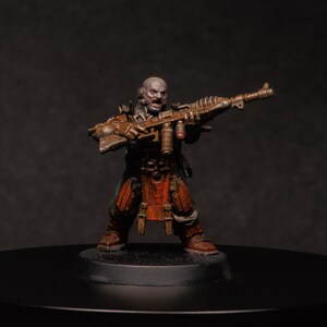 May include: A miniature figure of a bald warrior with a grimace, holding a detailed gun. The figure wears dark clothing with red and brown details, standing on a black base. The gun is a metallic brown colour.