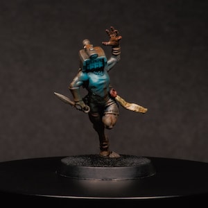 May include: A blue and brown painted miniature figurine of a female character wearing a helmet with a visor and holding a knife. The figurine is standing on a black base.