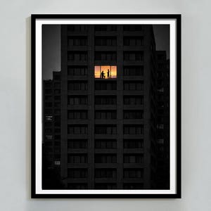May include: A framed art print featuring a high-rise building at dusk. The building's dark facade contrasts with a brightly lit window, silhouetting two figures. The warm sunset colors illuminate the window, creating a striking visual contrast.
