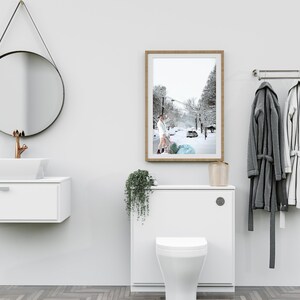 May include: A white bathroom with a white toilet, a white sink with a copper faucet, a round black mirror, a framed photograph of a snowy street, and a white towel rack with two towels hanging on it.