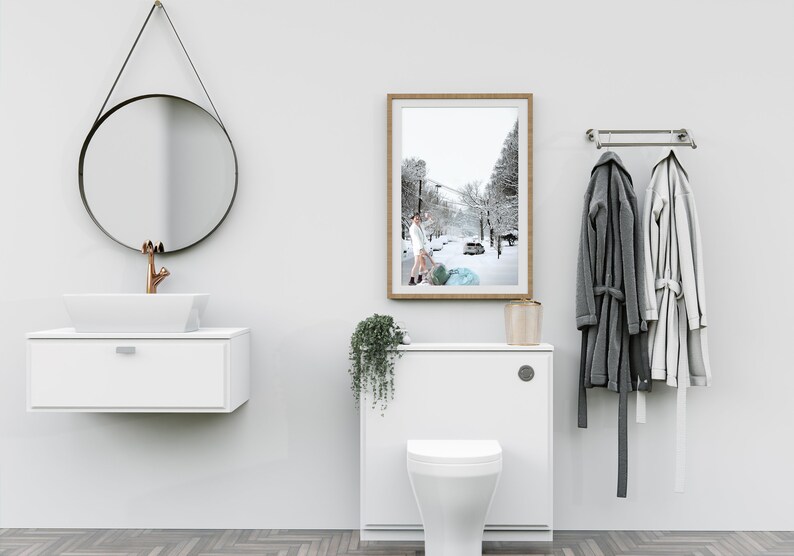 May include: A white bathroom with a white toilet, a white sink with a copper faucet, a round black mirror, a framed photograph of a snowy street, and a white towel rack with two towels hanging on it.