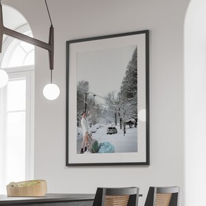 May include: A black framed photograph of a snowy street scene with a person in a white robe pulling a blue and white suitcase. The photograph is hanging on a white wall.