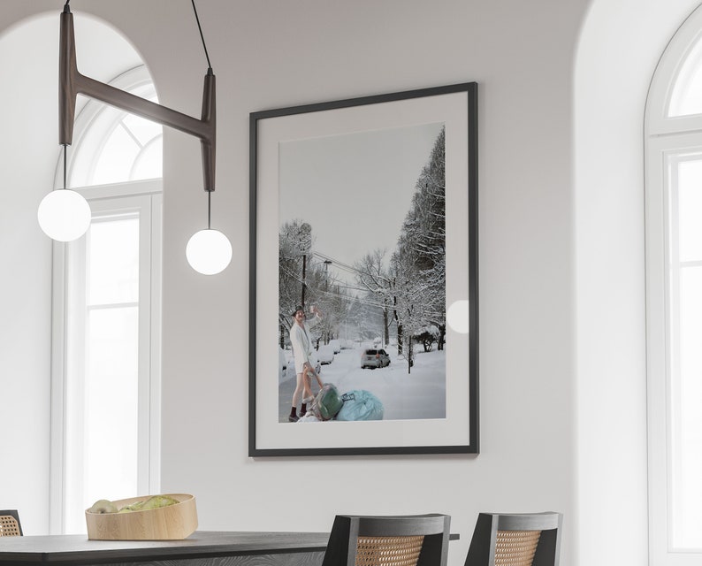 May include: A black framed photograph of a snowy street scene with a person in a white robe pulling a blue and white suitcase. The photograph is hanging on a white wall.