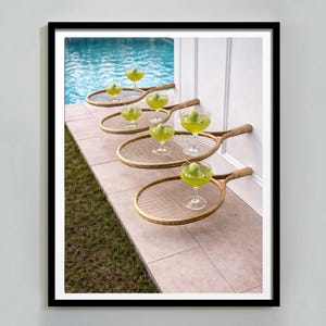 Può includere: Stampa incorniciata con una scena a bordo piscina con racchette da tennis che fungono da vassoi per bicchieri da cocktail. I cocktail sono verdi e le racchette sono disposte in fila. Lo sfondo comprende una piscina e un muro bianco.