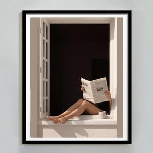 Puede incluir: Una impresión enmarcada muestra a una persona leyendo un periódico en una ventana. Las piernas de la persona son visibles, junto con una taza blanca. La ventana está abierta y el fondo es oscuro.