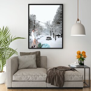 May include: A man in a white robe walks down a snowy street, pulling a large blue bag behind him. The street is lined with trees and houses, and the snow is falling softly.